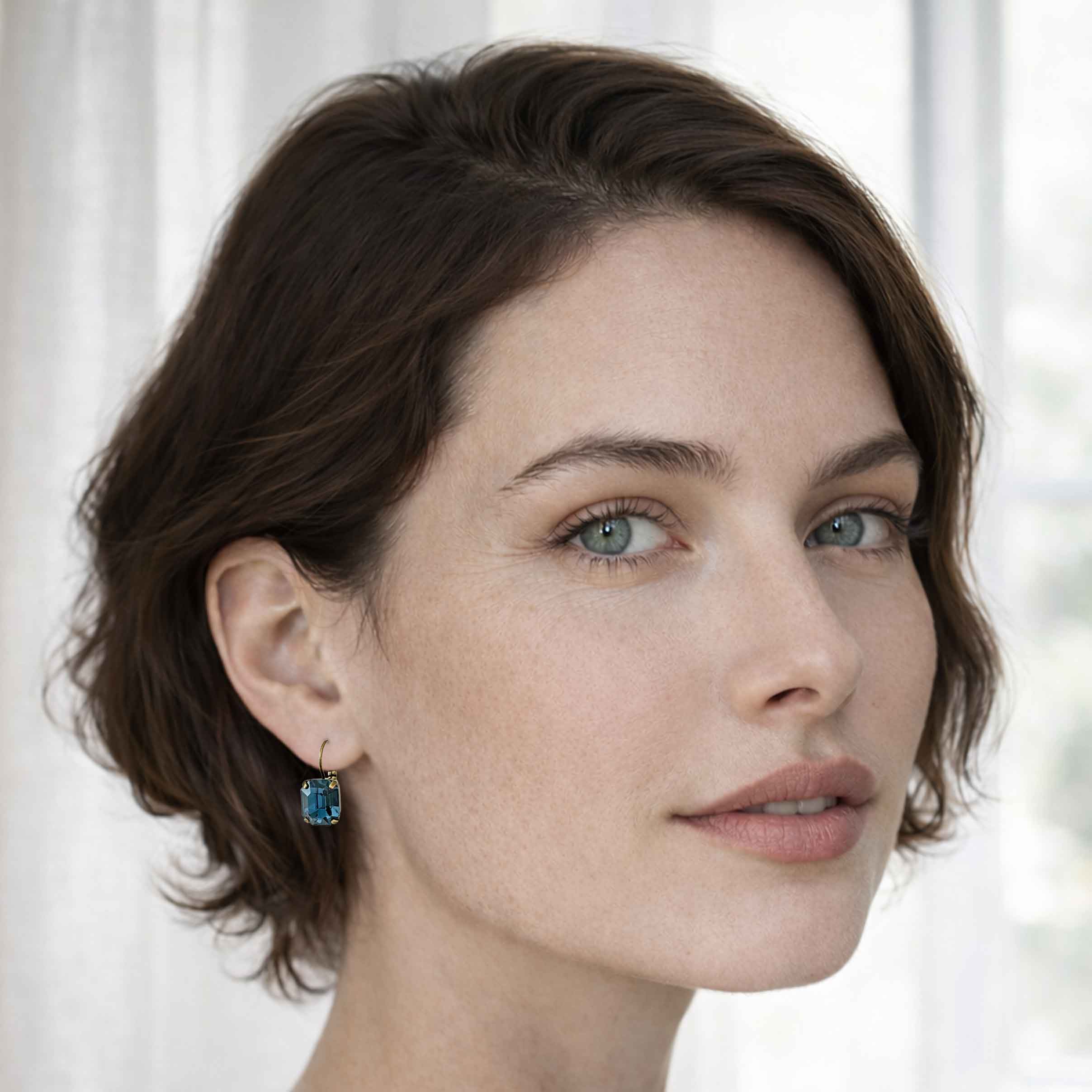 A young brunette wearing blue, emerald-cut crystal earrings by Petite Margaux.
