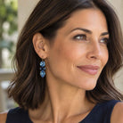 Brunette woman wearing light and dark blue crystal statement earrings by Petite Margaux with a navy dress.