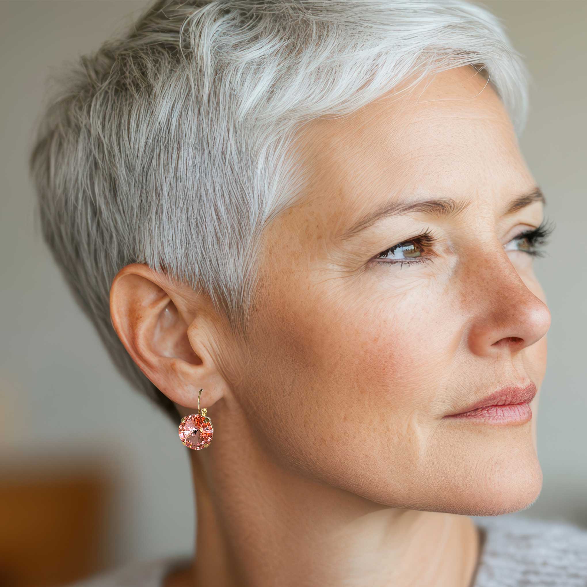 Woman with short gray hair wearing a round coral crystal drop earring against a neutral background.